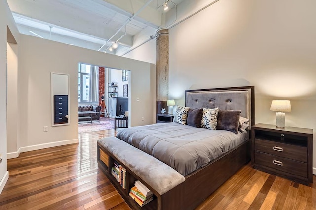 bedroom featuring baseboards, a towering ceiling, hardwood / wood-style flooring, beamed ceiling, and rail lighting