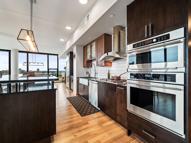 kitchen featuring a sink, backsplash, appliances with stainless steel finishes, wall chimney exhaust hood, and light wood finished floors