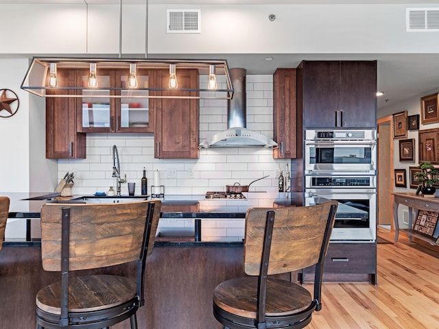 kitchen with a kitchen bar, wall chimney range hood, visible vents, and stainless steel double oven