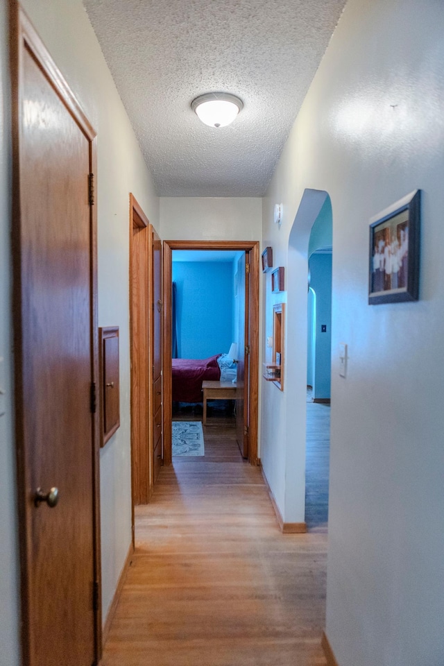 hall featuring light wood-type flooring, baseboards, arched walkways, and a textured ceiling