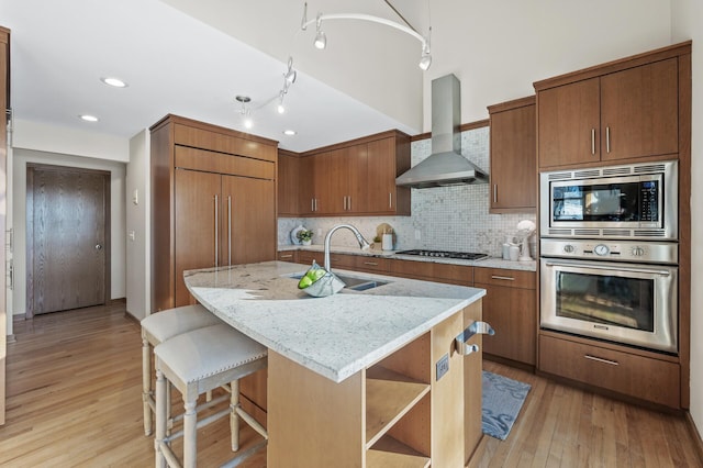 kitchen with wall chimney exhaust hood, brown cabinets, stainless steel appliances, light wood-type flooring, and a sink