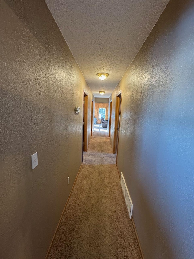 hallway featuring light colored carpet, visible vents, a textured wall, a textured ceiling, and baseboards