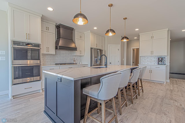 kitchen featuring white cabinets, an island with sink, appliances with stainless steel finishes, premium range hood, and a sink