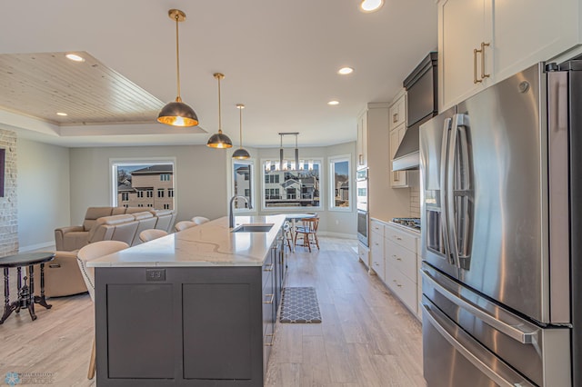 kitchen with light wood finished floors, a raised ceiling, a kitchen island with sink, stainless steel appliances, and a sink