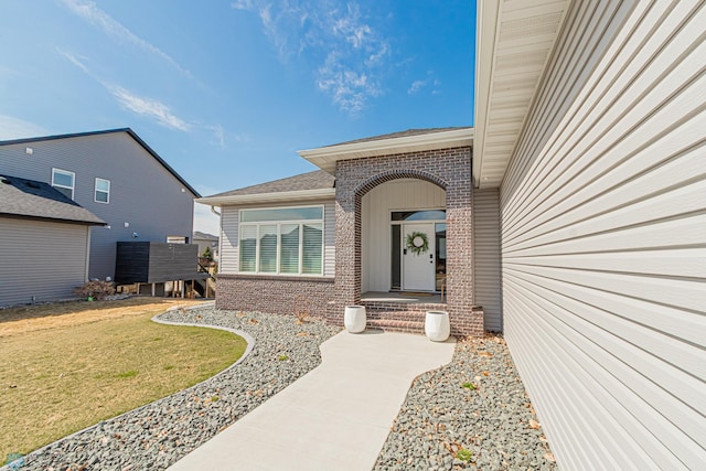 view of exterior entry featuring a lawn, a shingled roof, and brick siding