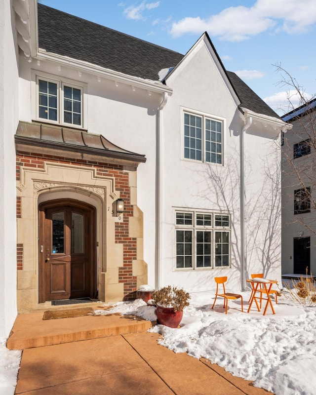 snow covered rear of property with a porch, roof with shingles, brick siding, and stucco siding