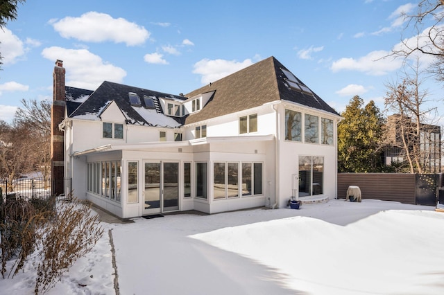 snow covered rear of property with a chimney, stucco siding, a shingled roof, a sunroom, and fence