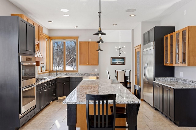 kitchen with a sink, light stone counters, a kitchen island, appliances with stainless steel finishes, and glass insert cabinets