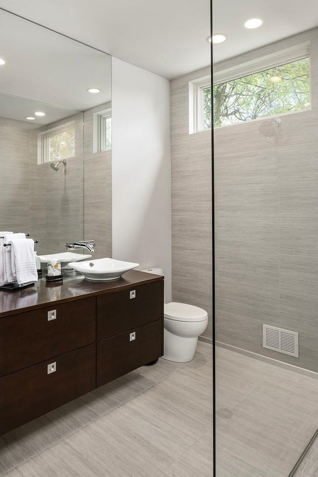 bathroom with toilet, vanity, visible vents, tile walls, and tiled shower