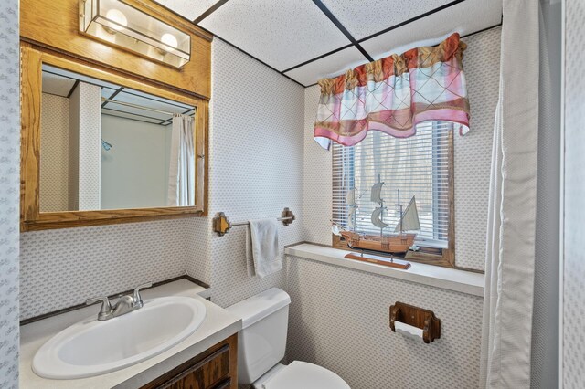 bathroom with a paneled ceiling, toilet, vanity, and wallpapered walls