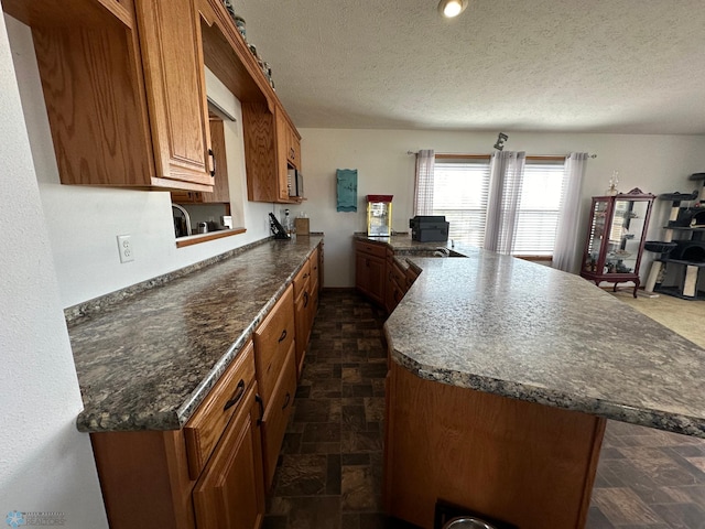 kitchen featuring a textured ceiling, a peninsula, stone finish floor, brown cabinetry, and dark countertops