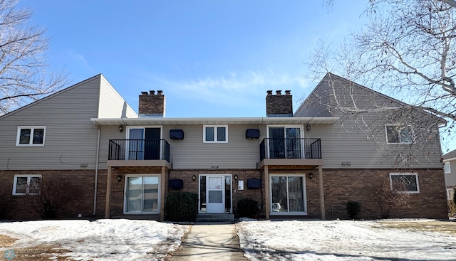 snow covered property featuring a balcony, a chimney, and brick siding