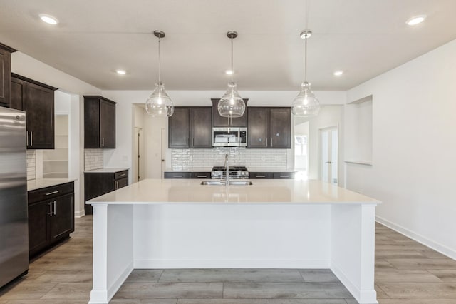 kitchen with light wood-style flooring, a kitchen island with sink, light countertops, dark brown cabinets, and appliances with stainless steel finishes