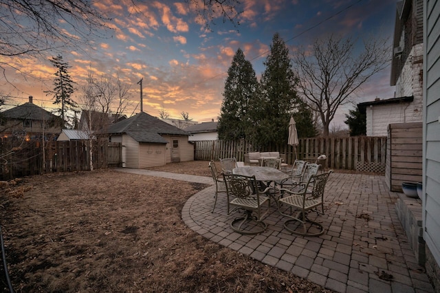 patio terrace at dusk featuring outdoor dining space, a fenced backyard, and an outbuilding