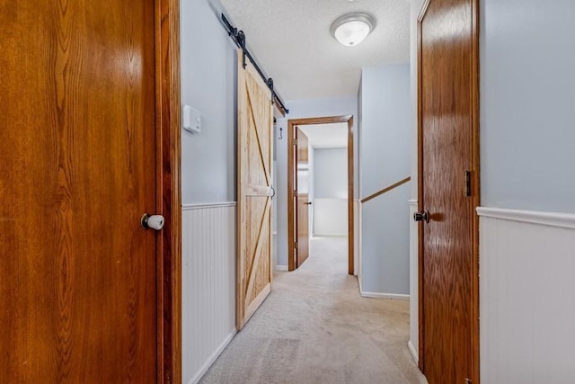 corridor with light carpet, a barn door, a wainscoted wall, and a textured ceiling