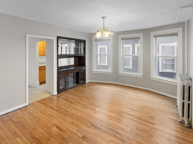 unfurnished dining area featuring radiator, an inviting chandelier, a textured ceiling, light wood-type flooring, and baseboards