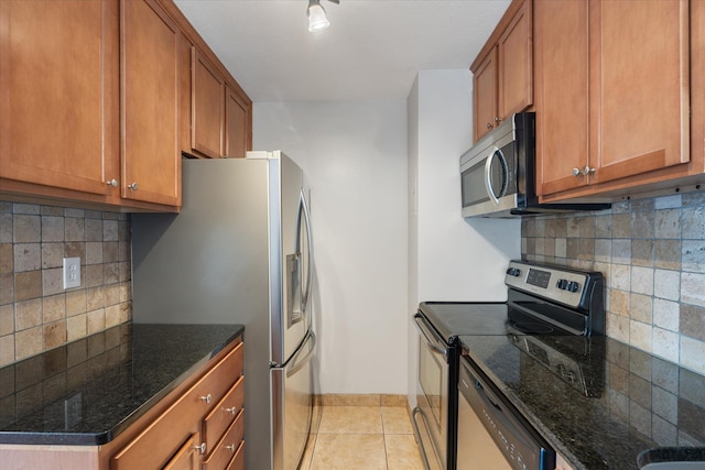 kitchen with light tile patterned floors, stainless steel appliances, brown cabinets, dark stone counters, and tasteful backsplash