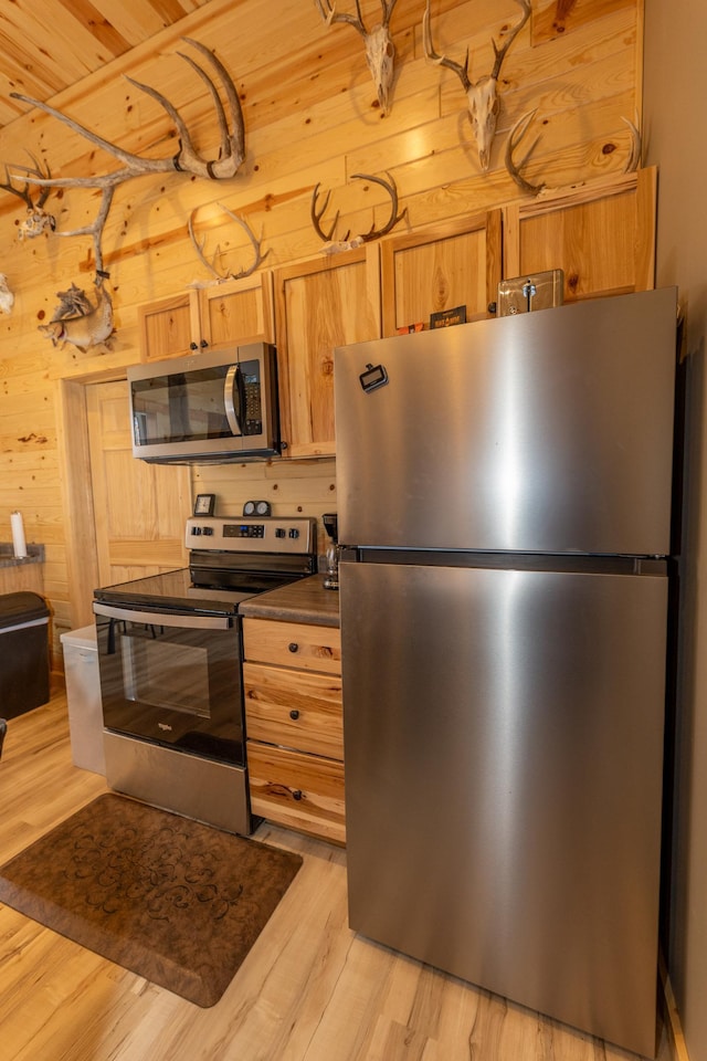 kitchen featuring dark countertops, light wood-style flooring, light brown cabinetry, appliances with stainless steel finishes, and wooden walls