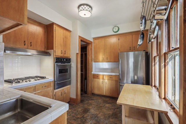 kitchen featuring tile countertops, appliances with stainless steel finishes, under cabinet range hood, backsplash, and a warming drawer