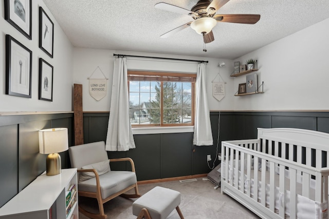 bedroom with a textured ceiling, a nursery area, ceiling fan, and wainscoting