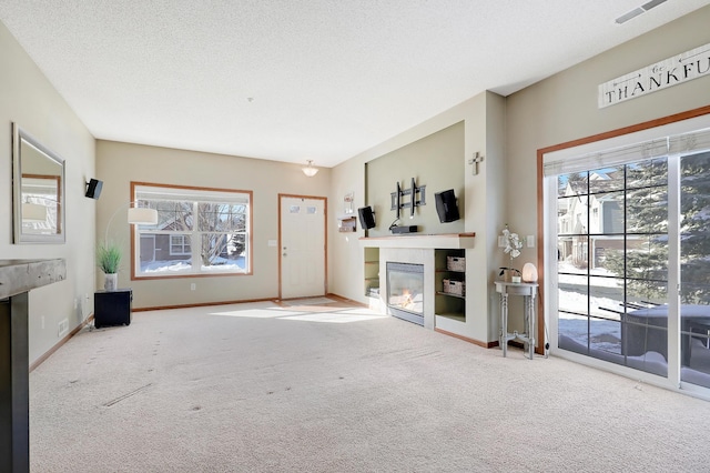 unfurnished living room featuring a textured ceiling, carpet floors, visible vents, baseboards, and a glass covered fireplace