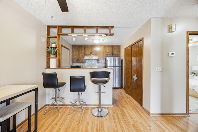 kitchen with a peninsula, freestanding refrigerator, under cabinet range hood, brown cabinets, and backsplash