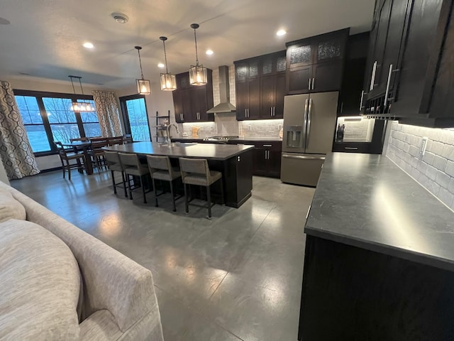 kitchen featuring a breakfast bar, a kitchen island with sink, wall chimney exhaust hood, stainless steel fridge with ice dispenser, and decorative backsplash