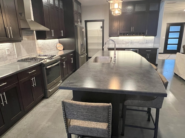 kitchen with an island with sink, a sink, appliances with stainless steel finishes, wall chimney range hood, and tasteful backsplash