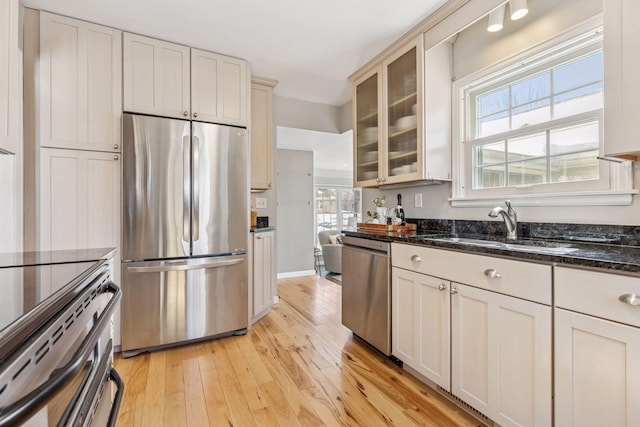 kitchen with glass insert cabinets, dark stone counters, light wood-type flooring, stainless steel appliances, and a sink