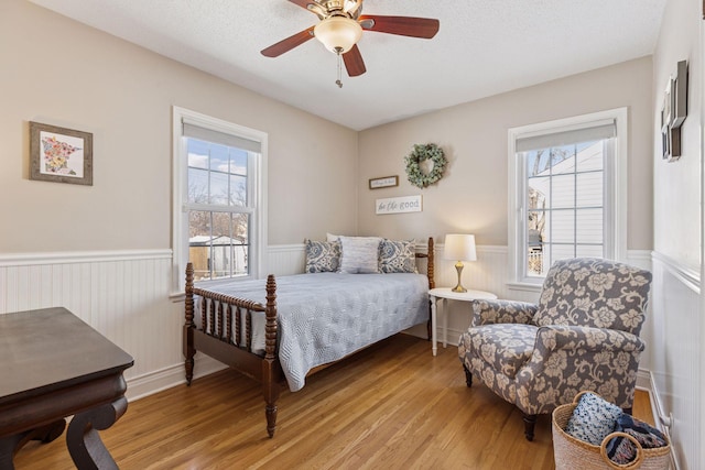 bedroom with a textured ceiling, ceiling fan, wainscoting, and light wood finished floors