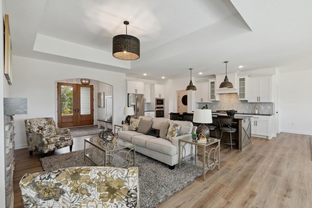 living room with baseboards, light wood-style flooring, french doors, arched walkways, and a raised ceiling