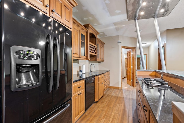 kitchen with brown cabinets, a sink, light wood-type flooring, dark stone counters, and black appliances