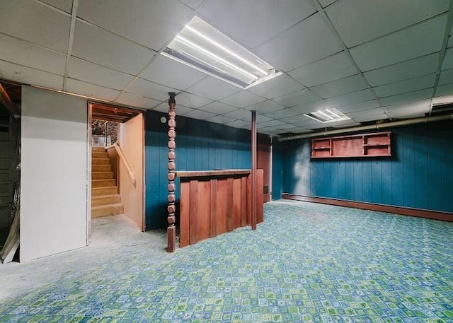 finished basement with a paneled ceiling, wooden walls, and stairs