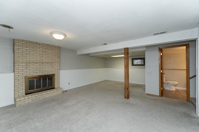 finished below grade area featuring visible vents, wainscoting, carpet flooring, a textured ceiling, and a brick fireplace