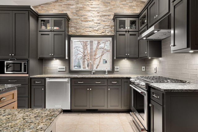 kitchen featuring dark stone counters, a sink, stainless steel appliances, under cabinet range hood, and backsplash