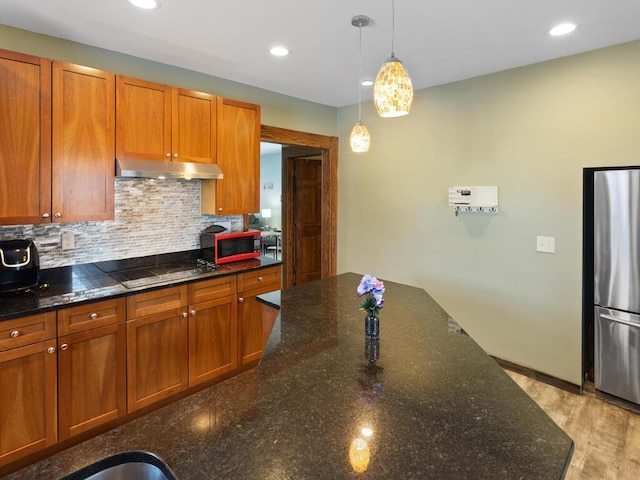 kitchen with decorative backsplash, brown cabinets, freestanding refrigerator, black electric stovetop, and under cabinet range hood