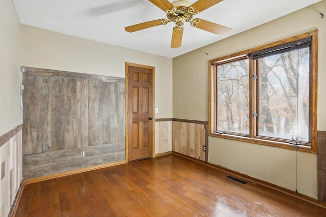 spare room with a textured ceiling, hardwood / wood-style flooring, visible vents, a ceiling fan, and wainscoting