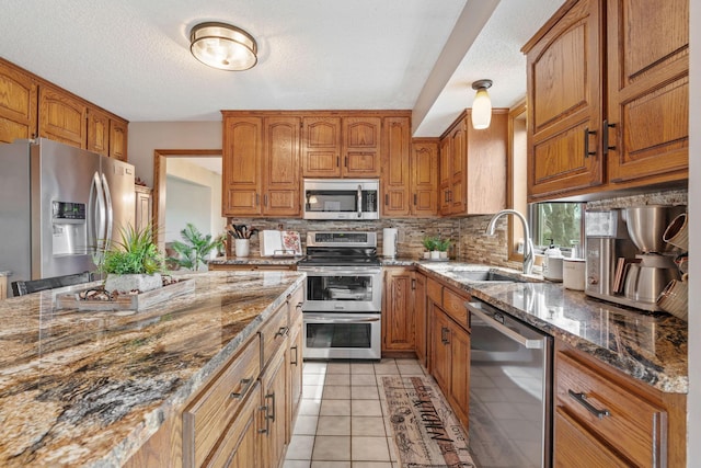 kitchen with brown cabinetry, a sink, dark stone countertops, backsplash, and stainless steel appliances