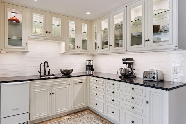 kitchen featuring a sink, decorative backsplash, glass insert cabinets, and white dishwasher