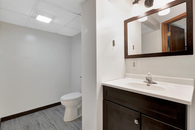 bathroom with toilet, a paneled ceiling, wood finished floors, vanity, and baseboards