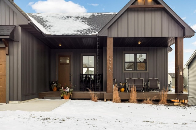 snow covered property entrance featuring roof with shingles, a porch, and board and batten siding