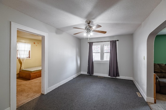 empty room featuring a ceiling fan, a textured ceiling, arched walkways, dark colored carpet, and baseboards