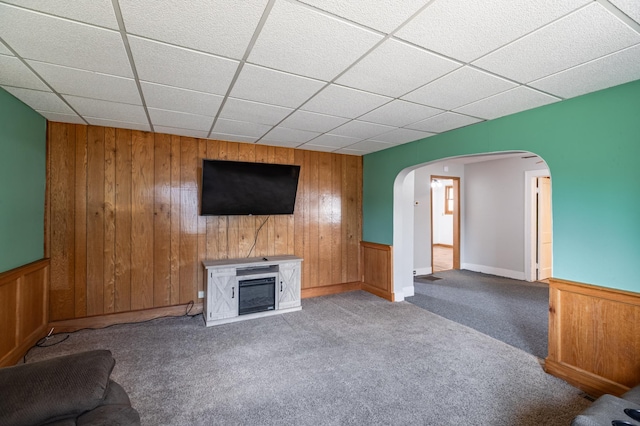 unfurnished living room featuring arched walkways, a drop ceiling, wood walls, and carpet floors