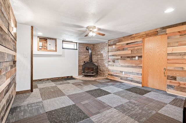 interior space featuring wooden walls, carpet flooring, baseboards, ceiling fan, and a wood stove