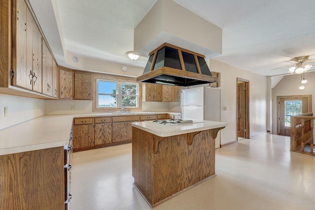 kitchen with a textured ceiling, white appliances, light countertops, wall chimney range hood, and brown cabinetry