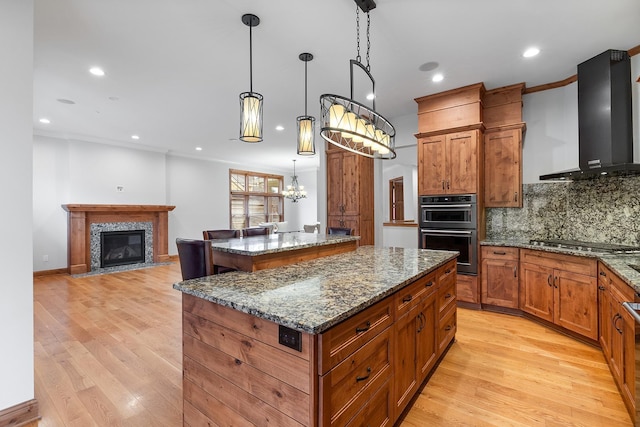 kitchen with brown cabinets, stainless steel gas cooktop, multiple ovens, a kitchen island, and wall chimney exhaust hood