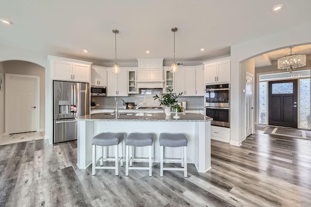 kitchen featuring arched walkways, appliances with stainless steel finishes, white cabinets, a sink, and an island with sink