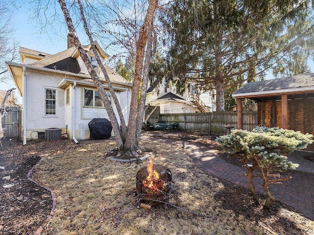 rear view of house featuring central AC unit, a fenced backyard, a shingled roof, stucco siding, and a fire pit