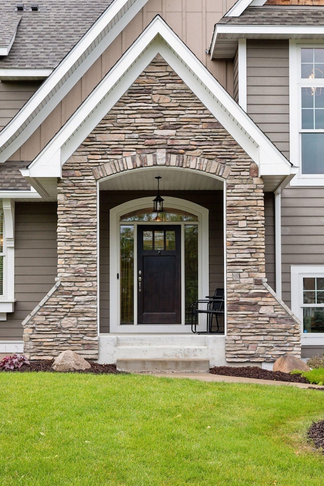 property entrance with stone siding, roof with shingles, and a lawn