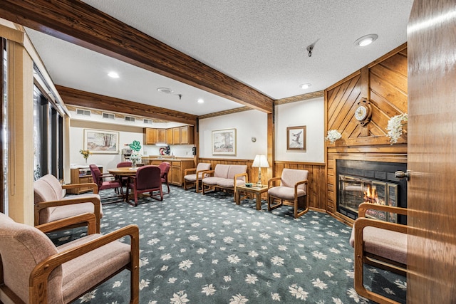 living area with wainscoting, a glass covered fireplace, beamed ceiling, a textured ceiling, and carpet flooring